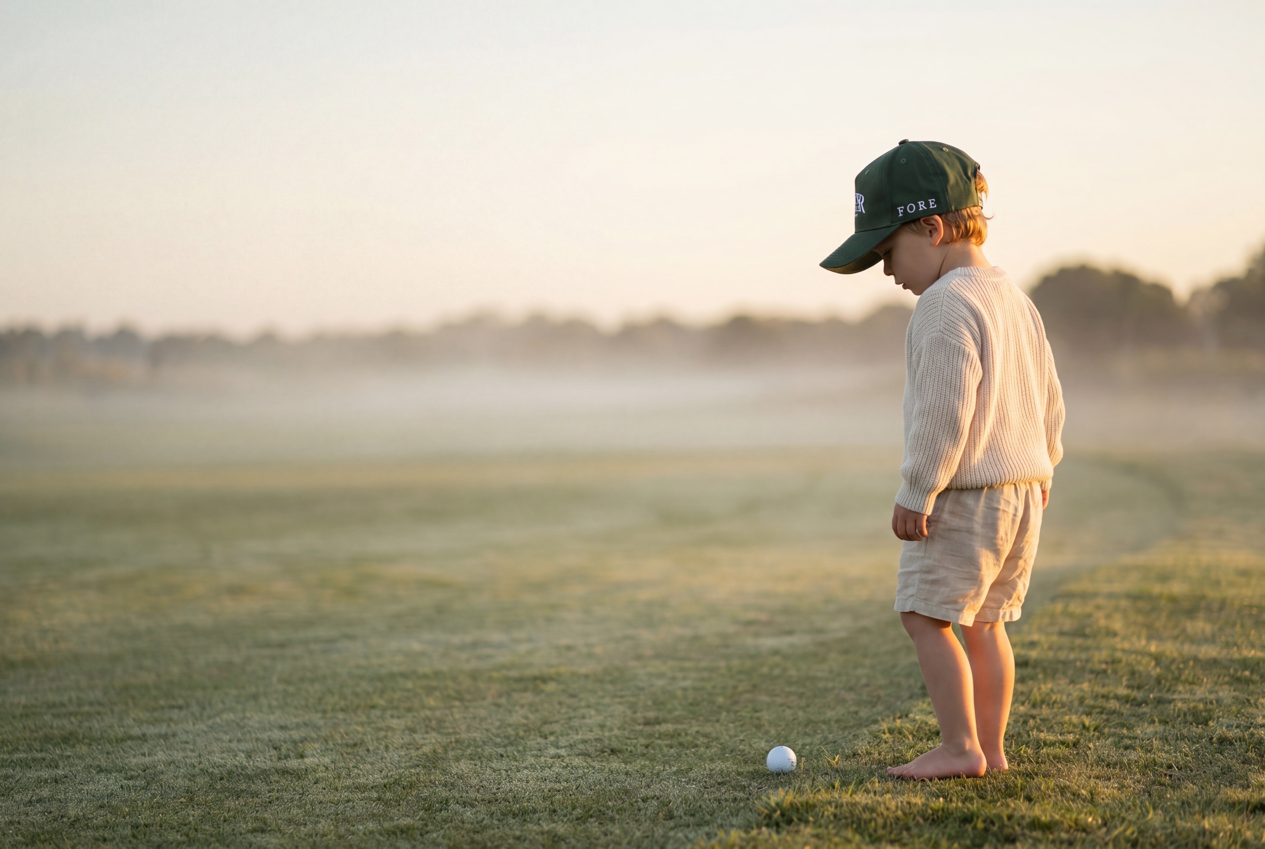 Toddler in green Bunker Boys hat standing barefoot on a misted fairway at sunrise with a single white golf ball at his feet.